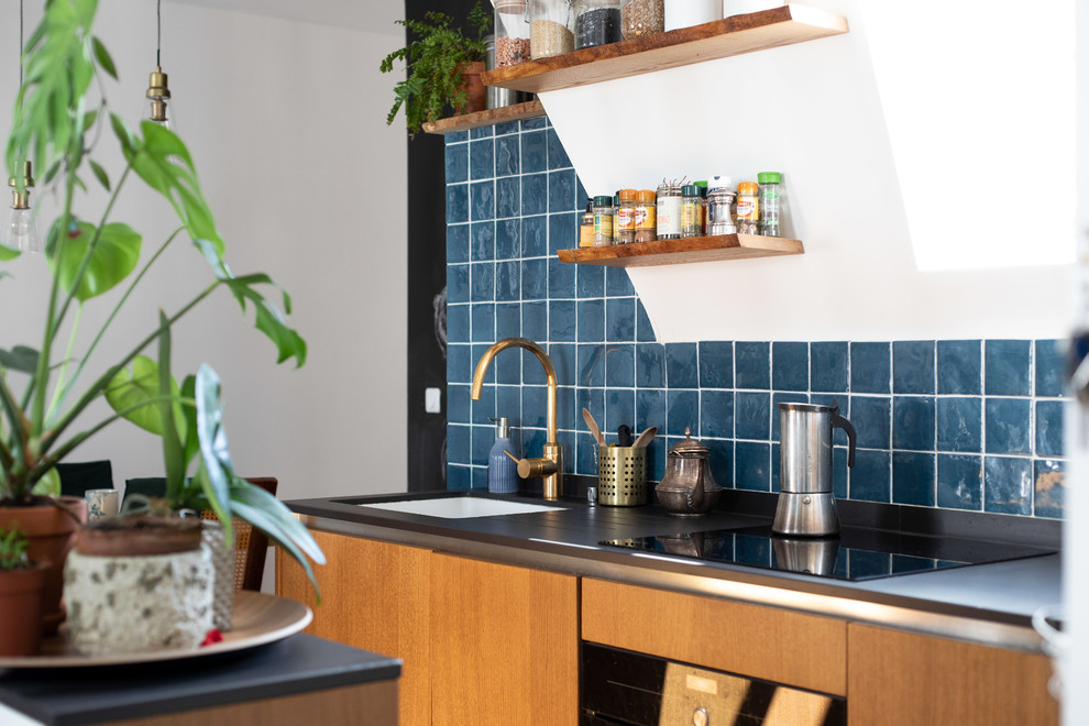 Photo of a small mediterranean galley open plan kitchen in Other with flat-panel cabinets, light wood cabinets, blue splashback, ceramic splashback, black appliances, ceramic floors, blue floor, black benchtop, an undermount sink, solid surface benchtops and a peninsula.