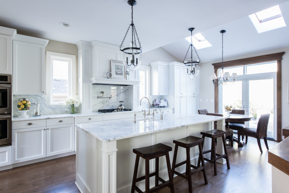 Photo of a large traditional single-wall open plan kitchen in Seattle with a farmhouse sink, recessed-panel cabinets, white cabinets, marble benchtops, white splashback, marble splashback, stainless steel appliances, dark hardwood floors, with island, brown floor and white benchtop.