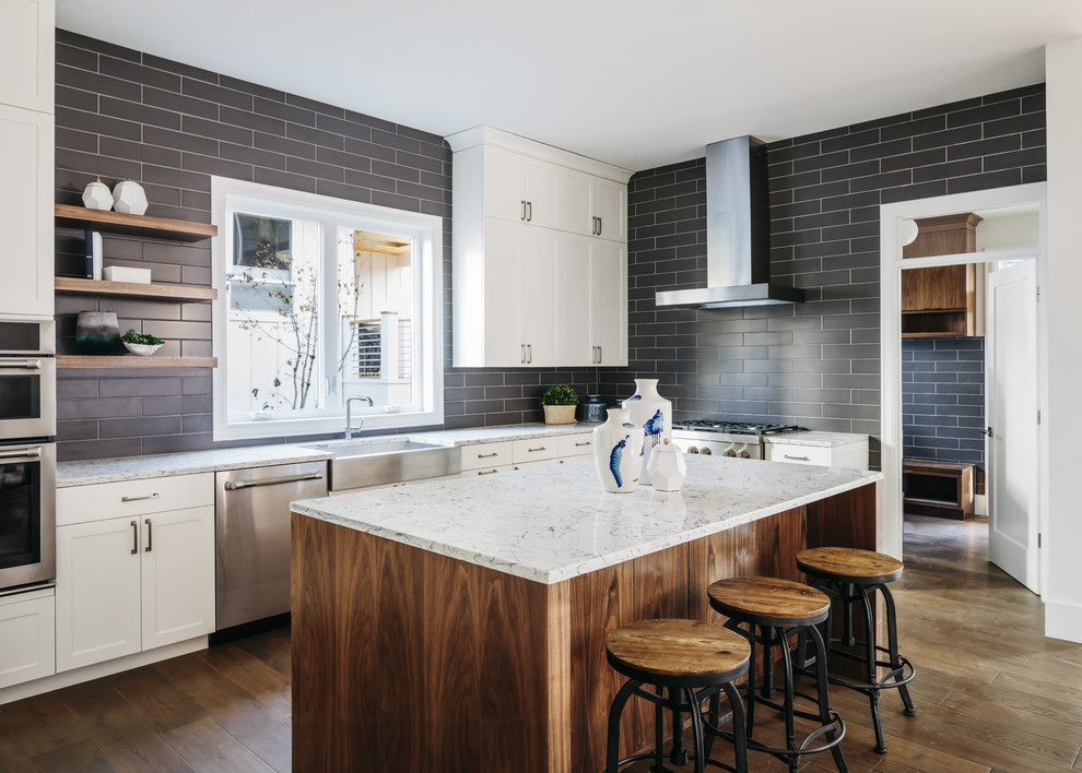 This is an example of a transitional l-shaped kitchen in San Francisco with a farmhouse sink, shaker cabinets, black splashback, subway tile splashback, stainless steel appliances, dark hardwood floors, with island, brown floor and multi-coloured benchtop.