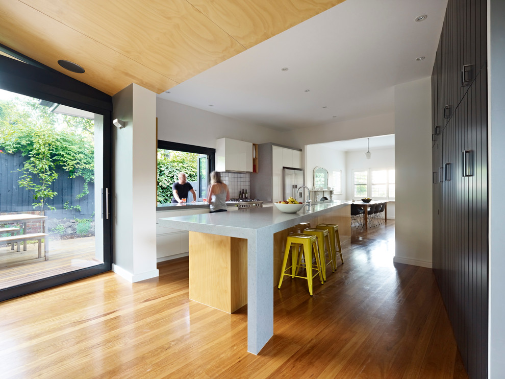 Photo of a large contemporary galley eat-in kitchen in Melbourne with an undermount sink, flat-panel cabinets, white cabinets, quartz benchtops, white splashback, porcelain splashback, stainless steel appliances, light hardwood floors and with island.