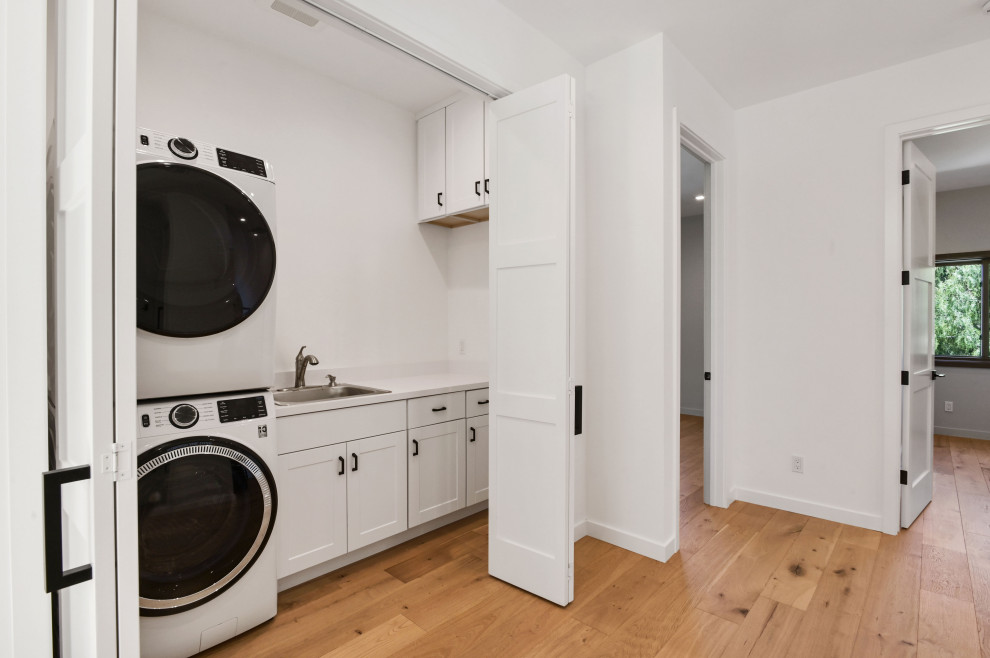 Design ideas for a mid-sized contemporary single-wall laundry cupboard in San Francisco with a single-bowl sink, shaker cabinets, white cabinets, quartz benchtops, white splashback, engineered quartz splashback, white walls, light hardwood floors, a stacked washer and dryer and white benchtop.
