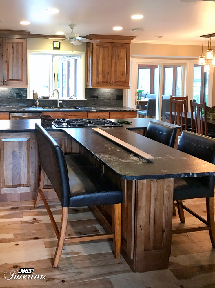Eat-in kitchen - large rustic l-shaped light wood floor and beige floor eat-in kitchen idea with an undermount sink, raised-panel cabinets, medium tone wood cabinets, granite countertops, gray backsplash, ceramic backsplash, stainless steel appliances and an island