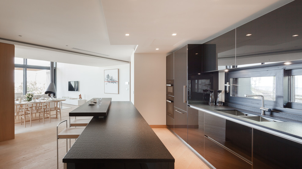 Contemporary separate kitchen in Other with flat-panel cabinets, light hardwood floors, with island, a double-bowl sink, black cabinets, black appliances, beige floor and black benchtop.
