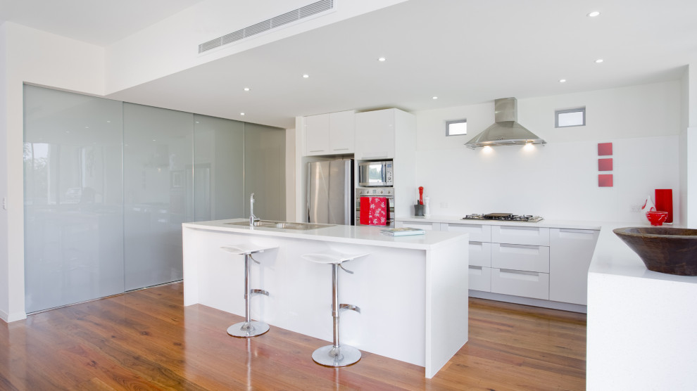 This is an example of an expansive modern l-shaped eat-in kitchen in Los Angeles with a drop-in sink, flat-panel cabinets, white cabinets, recycled glass benchtops, white splashback, stone slab splashback, coloured appliances, medium hardwood floors, with island, brown floor and white benchtop.