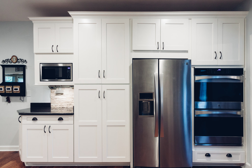 Example of a large transitional galley light wood floor and brown floor open concept kitchen design with an undermount sink, shaker cabinets, white cabinets, quartz countertops, white backsplash, stone tile backsplash, stainless steel appliances and a peninsula