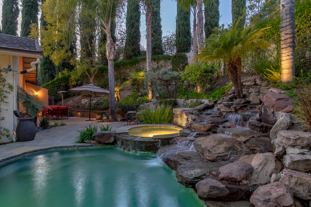 Large tropical backyard kidney-shaped natural pool in Los Angeles with a water feature and natural stone pavers.