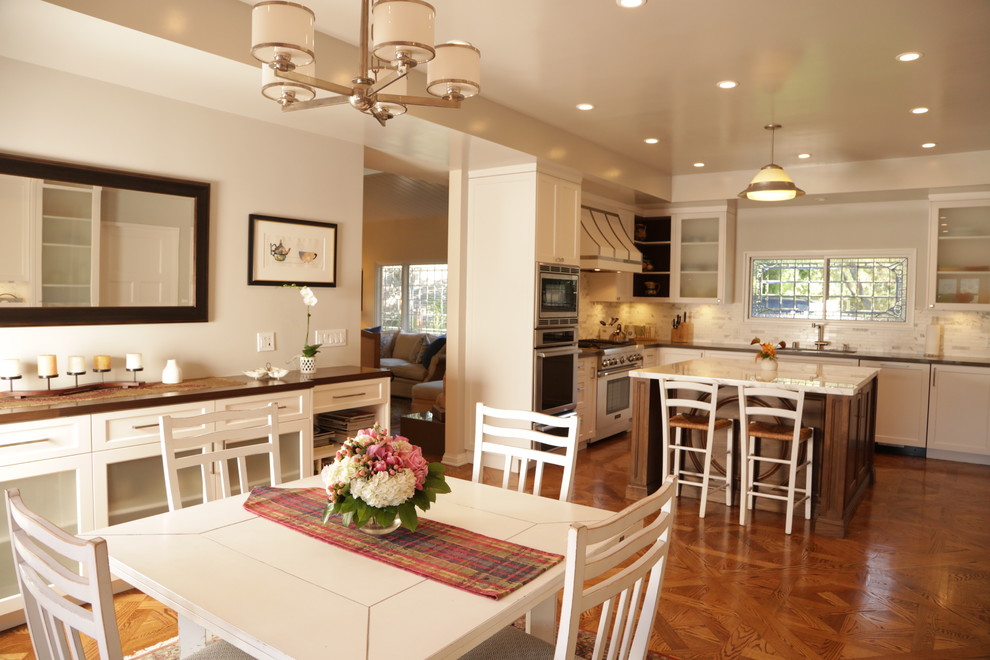 Inspiration for a large transitional u-shaped medium tone wood floor eat-in kitchen remodel in Los Angeles with an undermount sink, recessed-panel cabinets, white cabinets, wood countertops, white backsplash, stone tile backsplash, stainless steel appliances and an island
