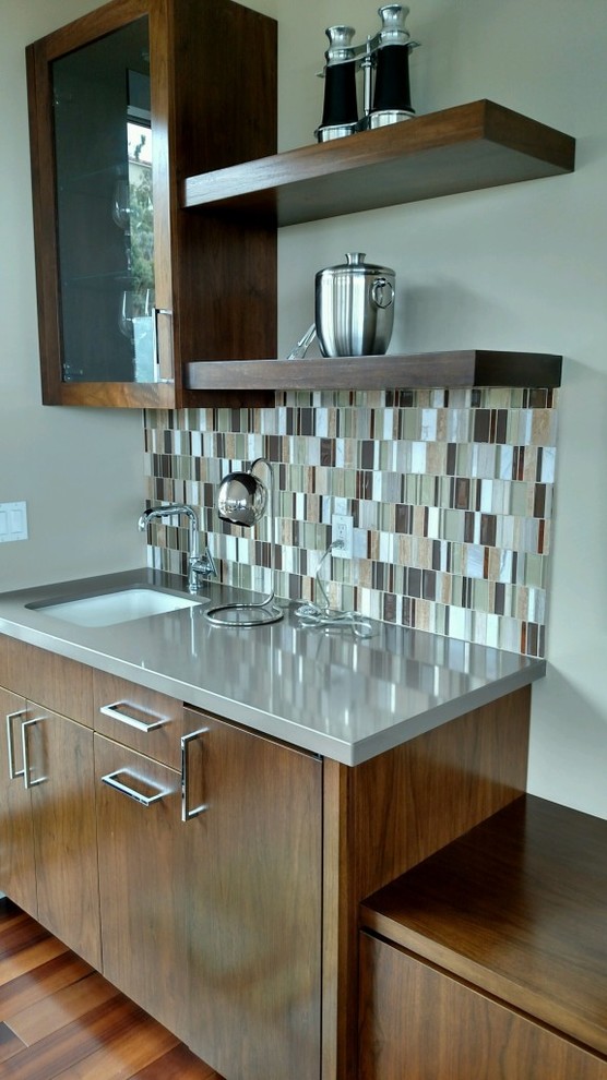 This is an example of a small modern single-wall wet bar in San Diego with an undermount sink, flat-panel cabinets, medium wood cabinets, solid surface benchtops, multi-coloured splashback, ceramic splashback, dark hardwood floors, brown floor and grey benchtop.