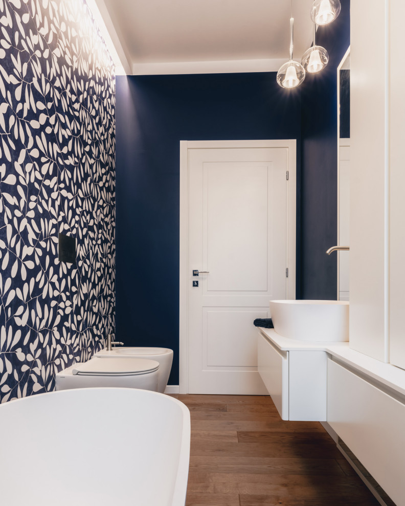 Foto på ett litet vit badrum, med släta luckor, blå skåp, ett fristående badkar, en toalettstol med separat cisternkåpa, flerfärgad kakel, porslinskakel, blå väggar, mörkt trägolv, ett fristående handfat, bänkskiva i akrylsten och brunt golv