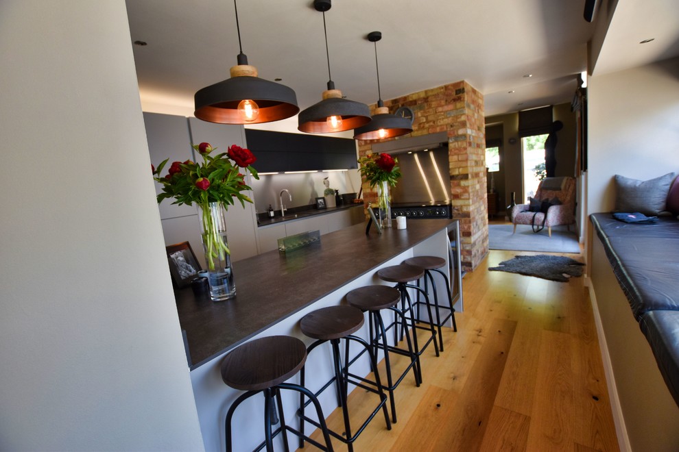 This is an example of a large contemporary eat-in kitchen in Cambridgeshire with an undermount sink, flat-panel cabinets, beige cabinets, black appliances, light hardwood floors, with island and grey benchtop.