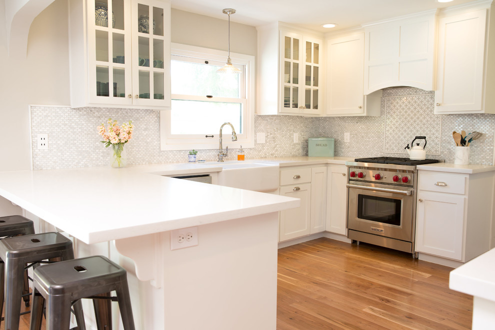 Example of a transitional medium tone wood floor kitchen design with a farmhouse sink, shaker cabinets, white cabinets, quartz countertops, white backsplash, stone tile backsplash and stainless steel appliances