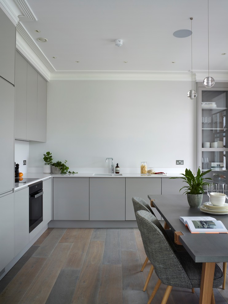 Design ideas for a large contemporary l-shaped eat-in kitchen in London with flat-panel cabinets, grey cabinets, no island, white benchtop, solid surface benchtops, a double-bowl sink, black appliances and medium hardwood floors.