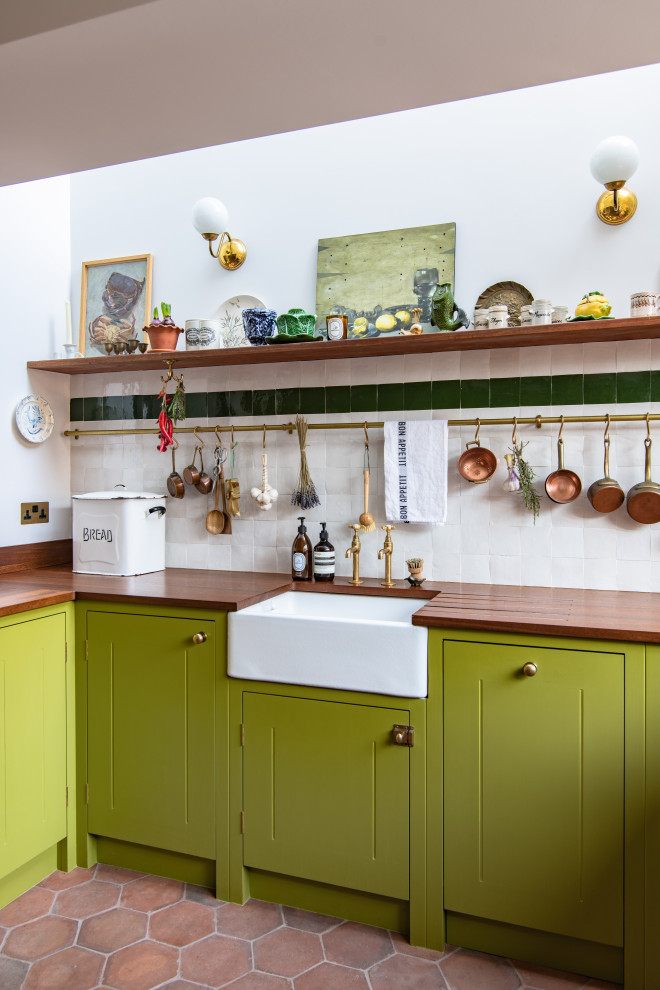 This is an example of a mid-sized contemporary u-shaped eat-in kitchen in London with a farmhouse sink, green cabinets, wood benchtops, white splashback, panelled appliances, terra-cotta floors, no island and brown benchtop.