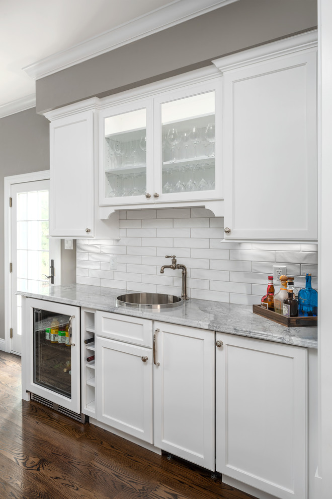Small traditional single-wall wet bar in St Louis with a drop-in sink, flat-panel cabinets, white cabinets, quartzite benchtops, white splashback, subway tile splashback, medium hardwood floors, brown floor and grey benchtop.