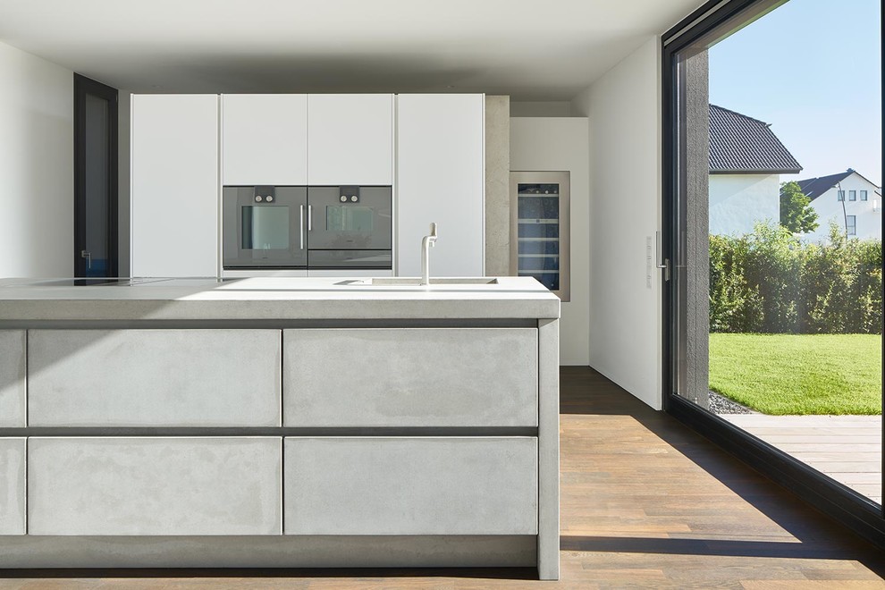 Contemporary single-wall kitchen in Berlin with a drop-in sink, flat-panel cabinets, grey cabinets, concrete benchtops, glass sheet splashback, medium hardwood floors and with island.