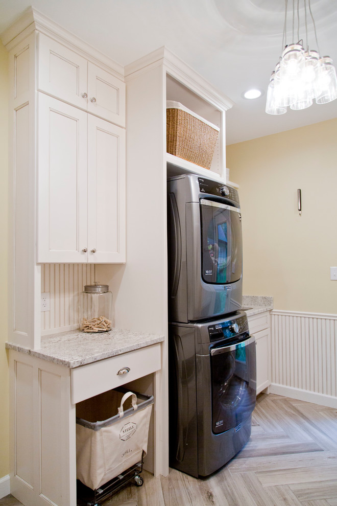 Photo of a mid-sized transitional single-wall utility room in St Louis with a drop-in sink, recessed-panel cabinets, white cabinets, granite benchtops, yellow walls, light hardwood floors and a stacked washer and dryer.