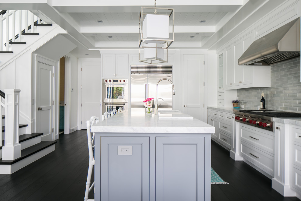 Large beach style l-shaped open plan kitchen in Orange County with a farmhouse sink, white cabinets, marble benchtops, grey splashback, ceramic splashback, stainless steel appliances, dark hardwood floors, with island and shaker cabinets.