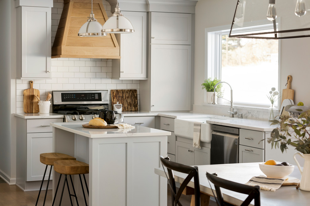Photo of a mid-sized country l-shaped eat-in kitchen in Minneapolis with a farmhouse sink, shaker cabinets, grey cabinets, quartz benchtops, white splashback, subway tile splashback, stainless steel appliances, medium hardwood floors, with island and white benchtop.