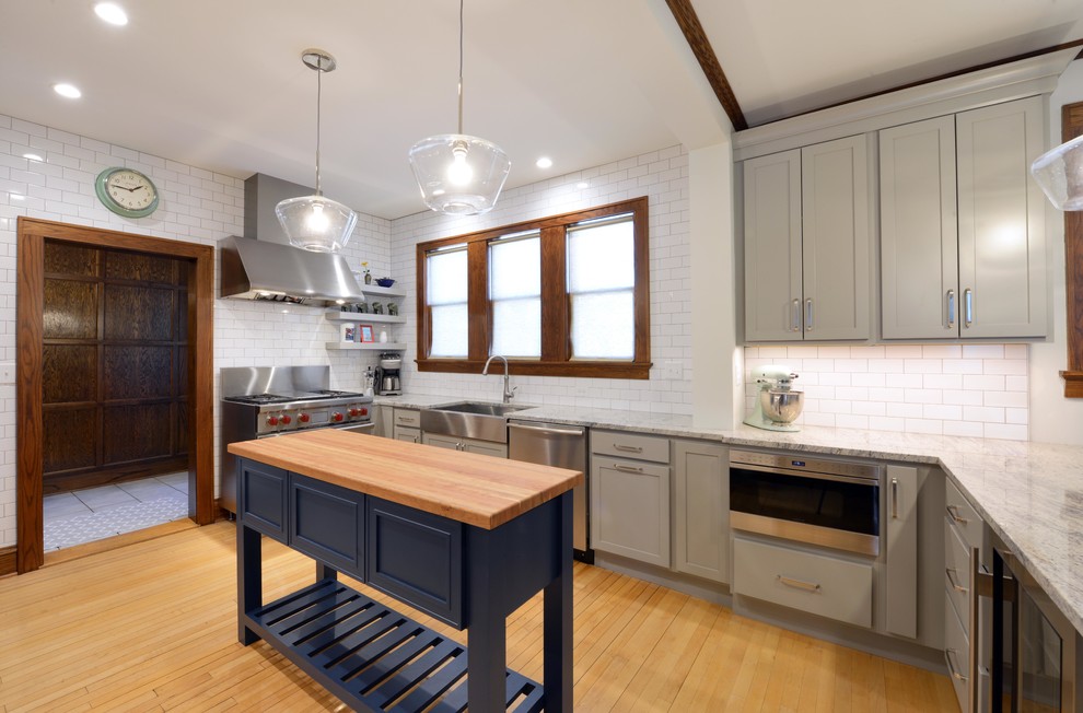 Example of a mid-sized transitional light wood floor and brown floor enclosed kitchen design with a farmhouse sink, shaker cabinets, gray cabinets, wood countertops, white backsplash, porcelain backsplash, stainless steel appliances, an island and brown countertops