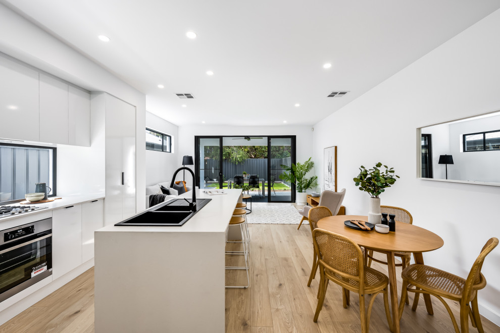 Small contemporary l-shaped open plan kitchen in Adelaide with a double-bowl sink, white cabinets, glass sheet splashback, black appliances, medium hardwood floors, with island, beige floor and white benchtop.