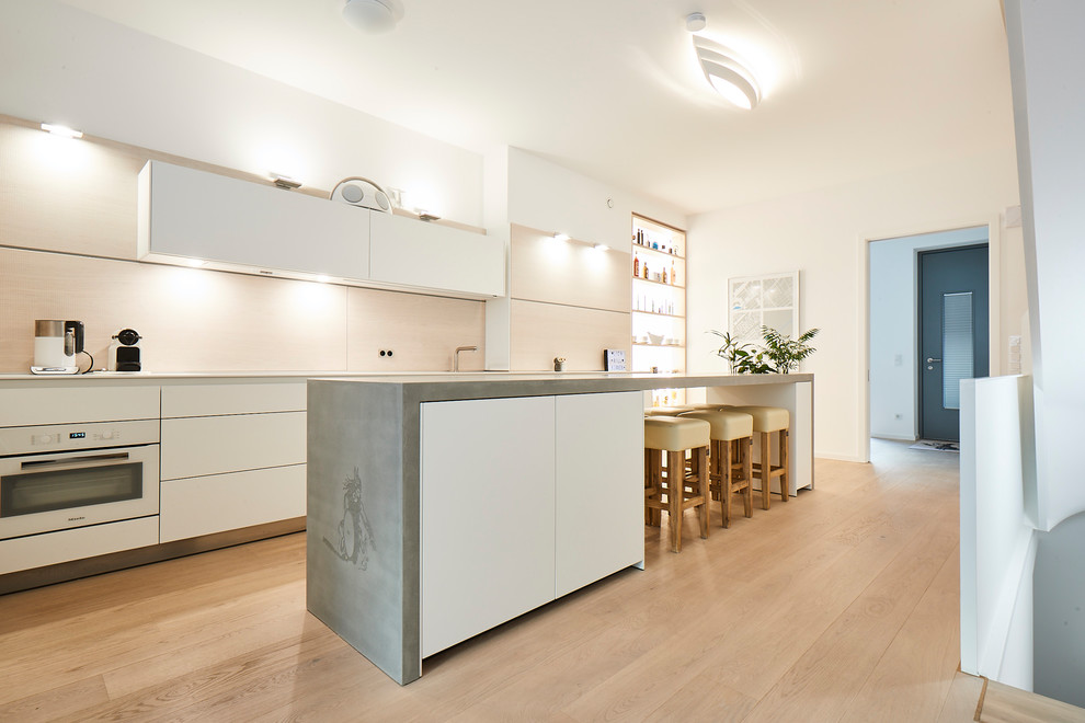Large modern galley open plan kitchen in Hamburg with an integrated sink, flat-panel cabinets, white cabinets, concrete benchtops, beige splashback, timber splashback, white appliances, medium hardwood floors, with island, brown floor and grey benchtop.