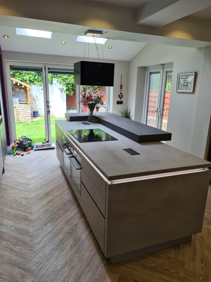 Large modern grey and purple single-wall kitchen/diner in Manchester with flat-panel cabinets, grey cabinets, composite countertops, metallic splashback, mirror splashback, black appliances, laminate floors, an island, brown floors, grey worktops and feature lighting.