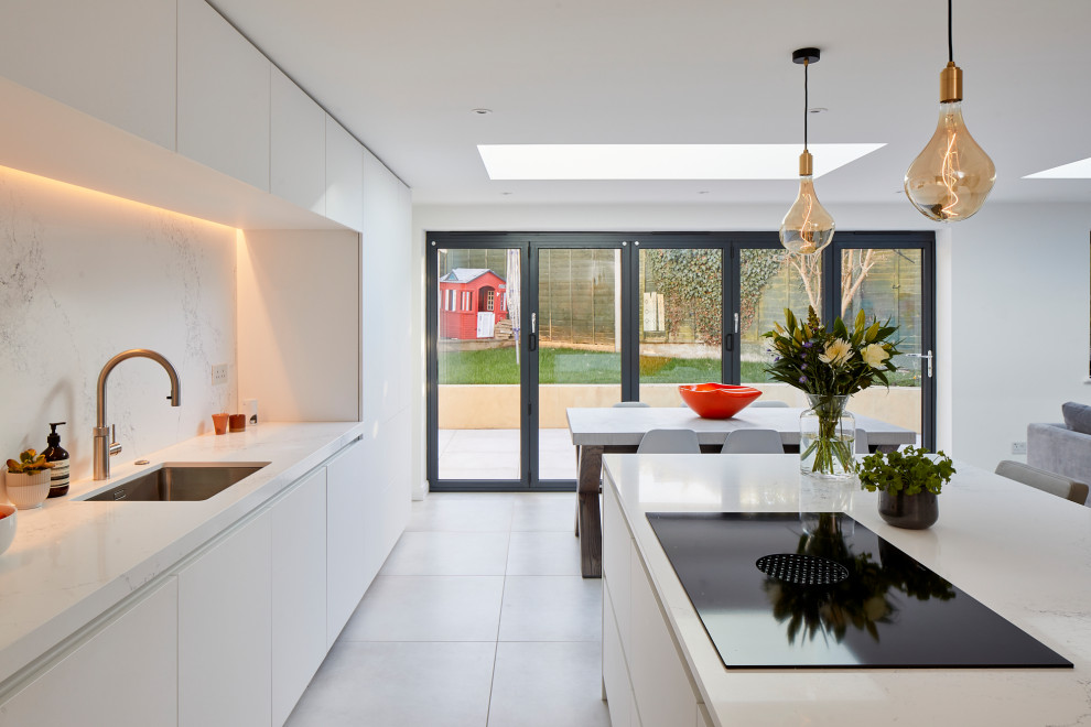 Photo of a large contemporary single-wall open plan kitchen in Kent with a submerged sink, flat-panel cabinets, white cabinets, engineered stone countertops, white splashback, engineered quartz splashback, stainless steel appliances, ceramic flooring, an island and white worktops.