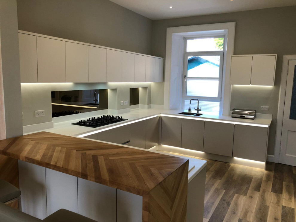 Photo of a large contemporary grey and white u-shaped open plan kitchen in Glasgow with an integrated sink, flat-panel cabinets, beige cabinets, quartz worktops, metallic splashback, mirror splashback, black appliances, vinyl flooring, a breakfast bar, brown floors, white worktops and feature lighting.