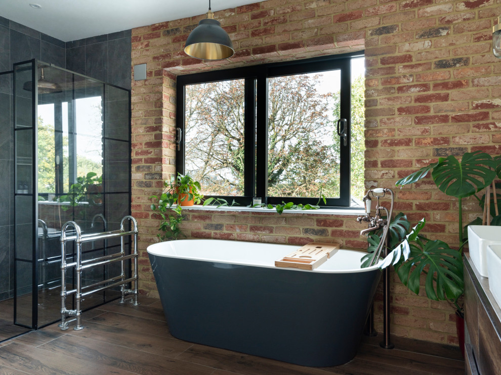 This is an example of an expansive contemporary kids bathroom in London with flat-panel cabinets, distressed cabinets, a freestanding tub, an open shower, a one-piece toilet, gray tile, ceramic tile, ceramic floors, a wall-mount sink, wood benchtops, an open shower, brown benchtops, a double vanity, a floating vanity and brick walls.