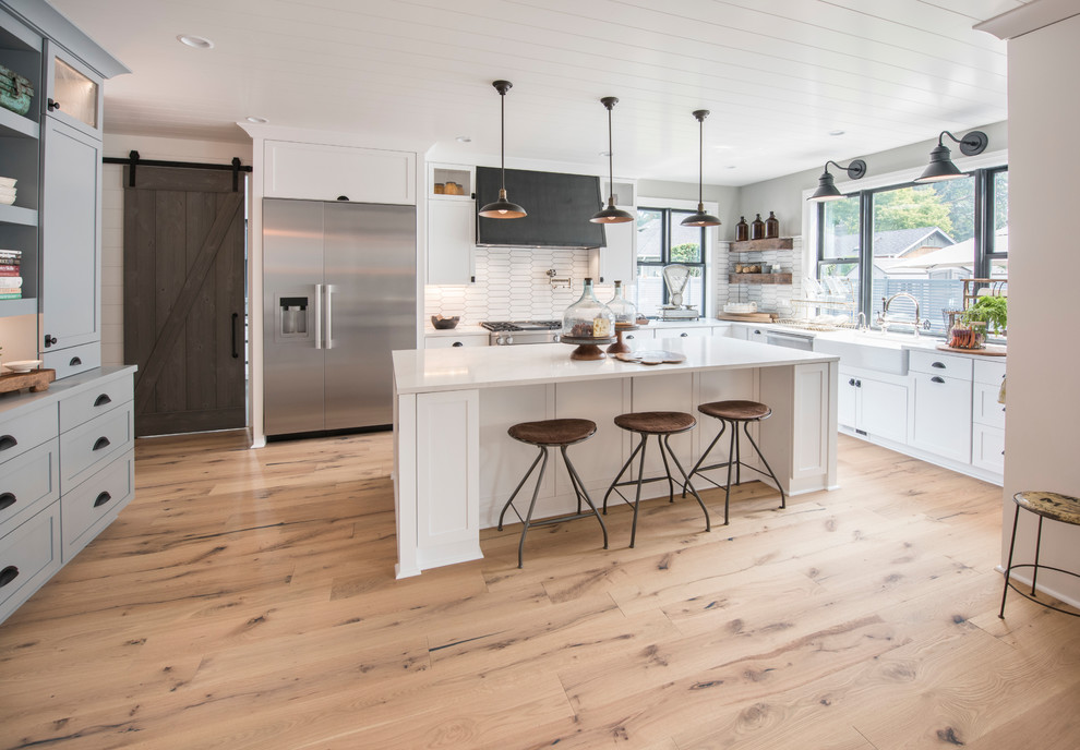 This is an example of a country l-shaped kitchen in Seattle with a farmhouse sink, shaker cabinets, white cabinets, white splashback, stainless steel appliances, light hardwood floors, with island, beige floor and white benchtop.