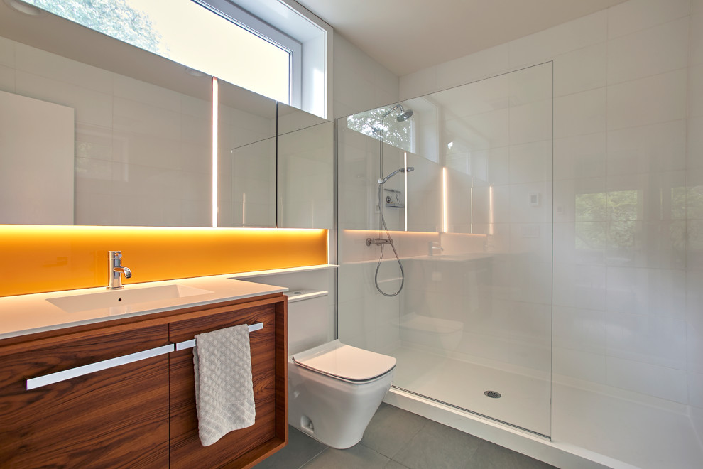 Photo of a mid-sized modern 3/4 bathroom in New York with flat-panel cabinets, medium wood cabinets, an alcove shower, an urinal, white tile, glass tile, white walls, porcelain floors, an integrated sink, solid surface benchtops, grey floor, an open shower and white benchtops.