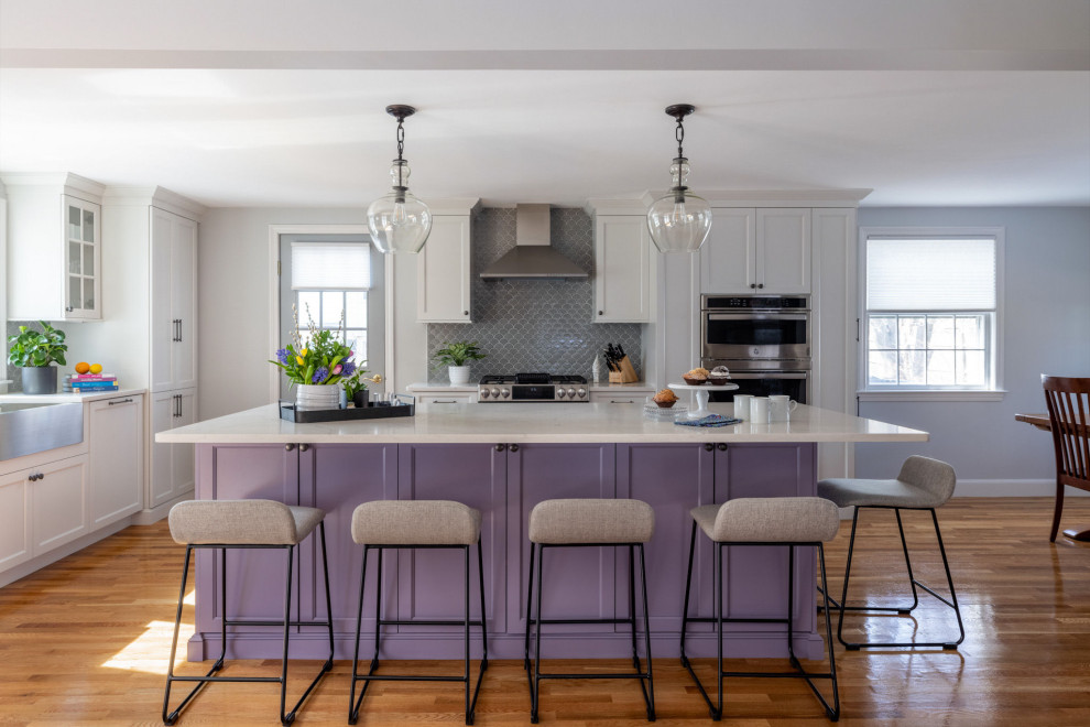 Photo of a traditional l-shaped kitchen in Boston with a belfast sink, shaker cabinets, purple cabinets, engineered stone countertops, grey splashback, ceramic splashback, stainless steel appliances, light hardwood flooring, an island and white worktops.