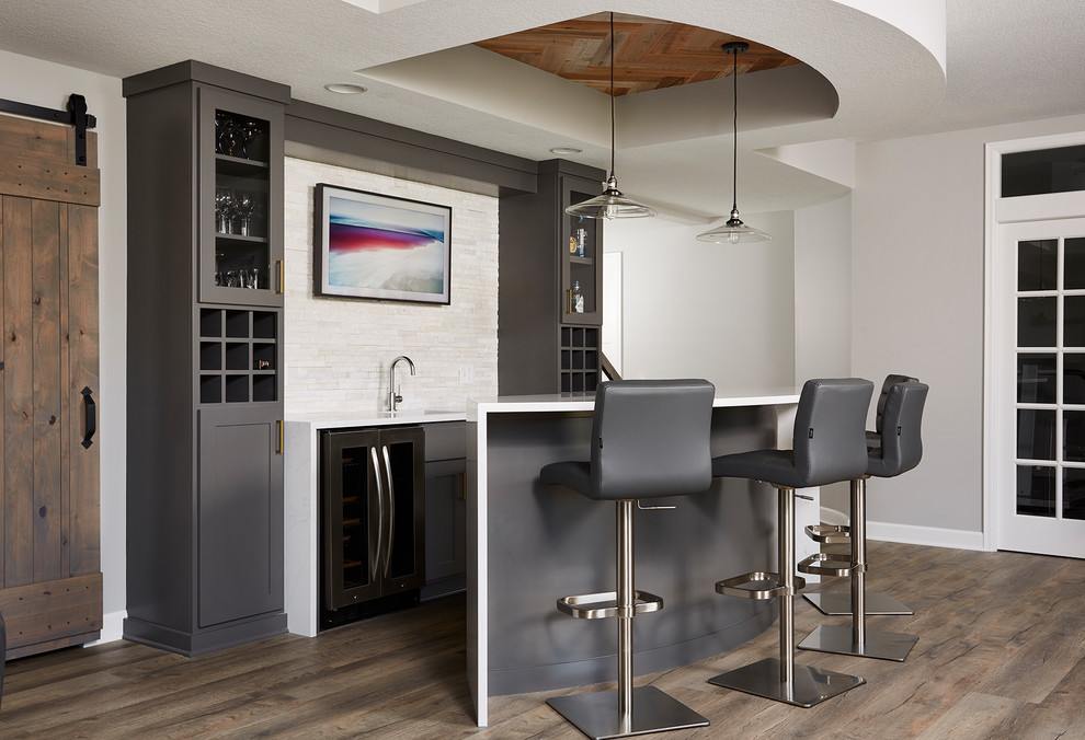 This is an example of a transitional galley wet bar in Minneapolis with an undermount sink, shaker cabinets, grey cabinets, beige splashback, matchstick tile splashback, medium hardwood floors, brown floor and white benchtop.