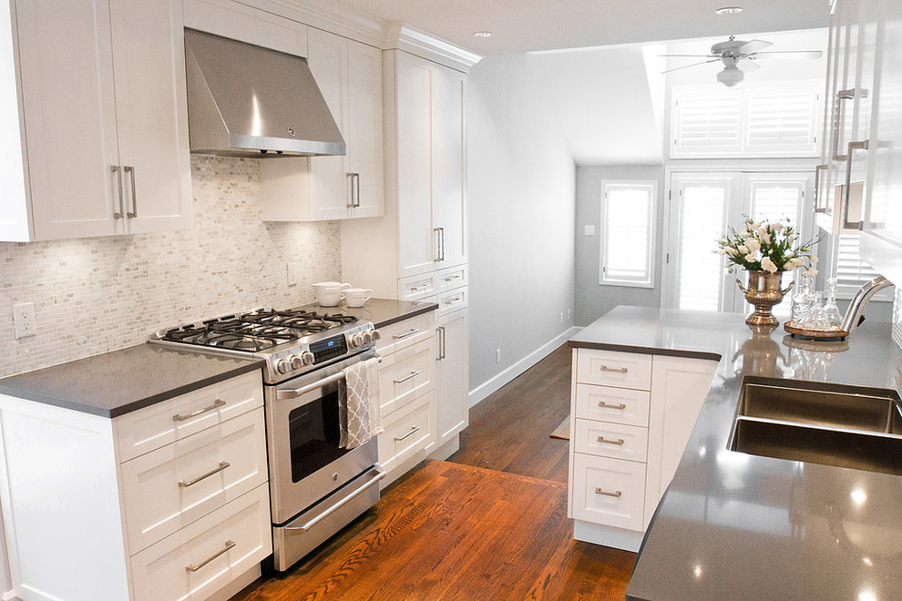 This is an example of a mid-sized transitional galley open plan kitchen in Vancouver with a double-bowl sink, shaker cabinets, white cabinets, quartz benchtops, multi-coloured splashback, stone tile splashback, stainless steel appliances, medium hardwood floors and a peninsula.
