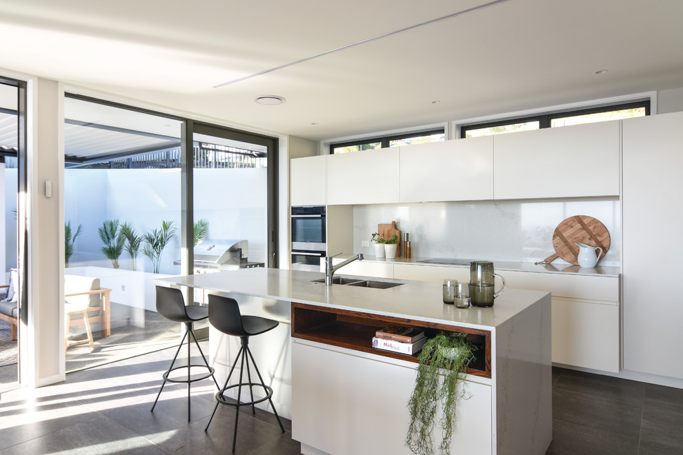 Photo of a mid-sized contemporary single-wall kitchen in Christchurch with a double-bowl sink, flat-panel cabinets, white cabinets, white splashback, stainless steel appliances, with island, grey floor and white benchtop.
