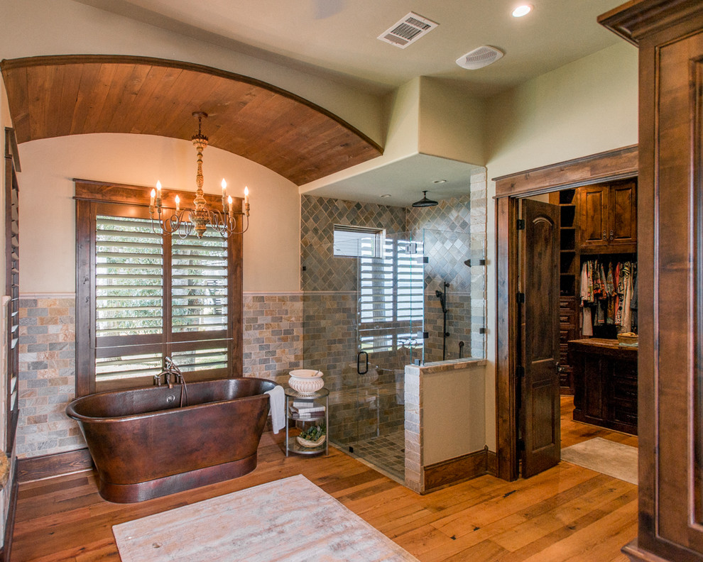 This is an example of a large country master bathroom in Houston with recessed-panel cabinets, distressed cabinets, a freestanding tub, a corner shower, beige tile, stone tile, beige walls, medium hardwood floors, an undermount sink, granite benchtops, brown floor and a hinged shower door.