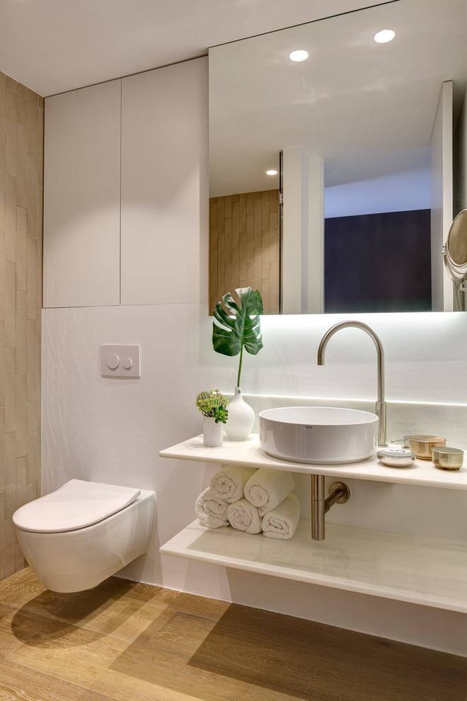 This is an example of a beach style 3/4 bathroom in Alicante-Costa Blanca with white cabinets, medium hardwood floors, a vessel sink, marble benchtops, white benchtops, an alcove shower, a wall-mount toilet, white walls, beige floor and a hinged shower door.