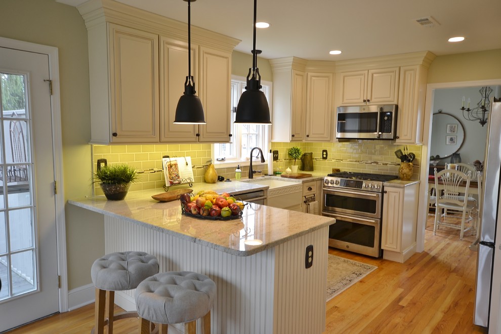 Photo of a mid-sized traditional l-shaped eat-in kitchen in Philadelphia with a farmhouse sink, raised-panel cabinets, white cabinets, granite benchtops, beige splashback, ceramic splashback, stainless steel appliances, light hardwood floors, a peninsula and brown floor.