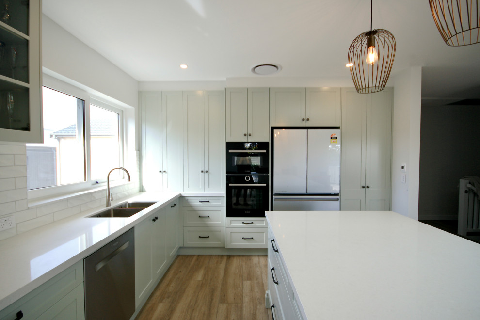 Photo of a large nautical l-shaped open plan kitchen in Sydney with a submerged sink, shaker cabinets, green cabinets, engineered stone countertops, white splashback, metro tiled splashback, black appliances, medium hardwood flooring, an island, brown floors and white worktops.