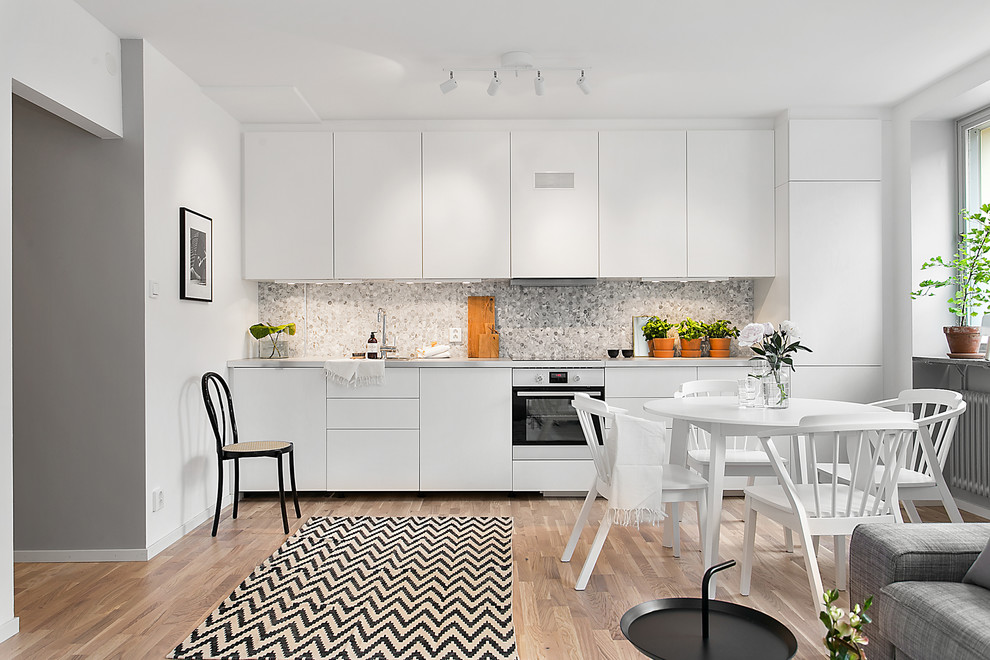 Photo of a mid-sized scandinavian single-wall open plan kitchen in Stockholm with flat-panel cabinets, white cabinets, grey splashback, white appliances, medium hardwood floors and no island.