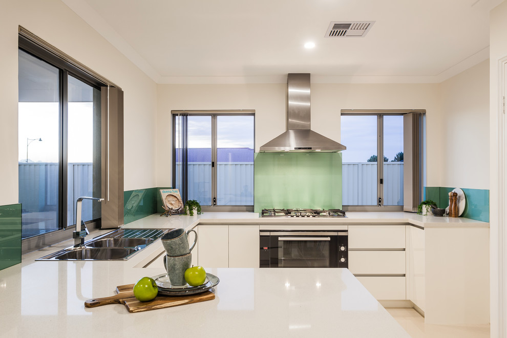 Inspiration for a contemporary u-shaped eat-in kitchen in Perth with a drop-in sink, flat-panel cabinets, white cabinets, blue splashback, glass sheet splashback, black appliances, a peninsula, beige floor and white benchtop.