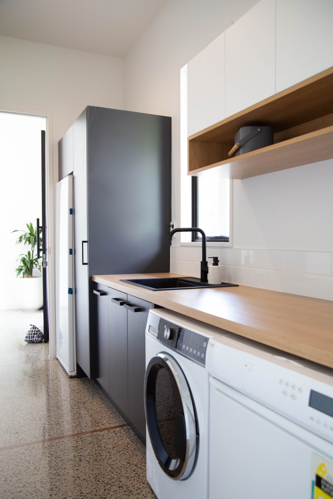 Photo of a mid-sized modern single-wall utility room in Other with a drop-in sink, flat-panel cabinets, black cabinets, laminate benchtops, white walls, concrete floors and a side-by-side washer and dryer.