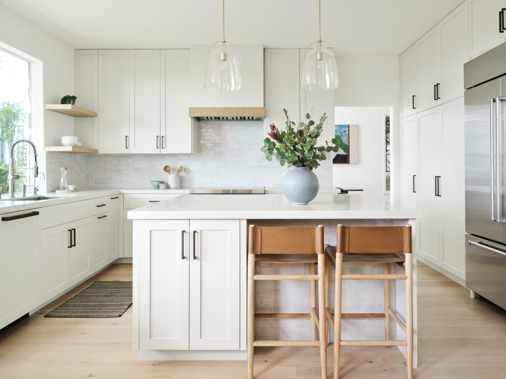 Photo of a beach style u-shaped kitchen in Los Angeles with an undermount sink, shaker cabinets, white cabinets, white splashback, stainless steel appliances, light hardwood floors, with island and white benchtop.