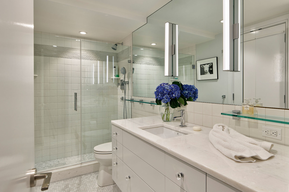 Photo of a small modern bathroom in Philadelphia with an alcove shower, a one-piece toilet, white tile, white walls, ceramic floors, an undermount sink, marble benchtops, grey floor, a hinged shower door and white benchtops.