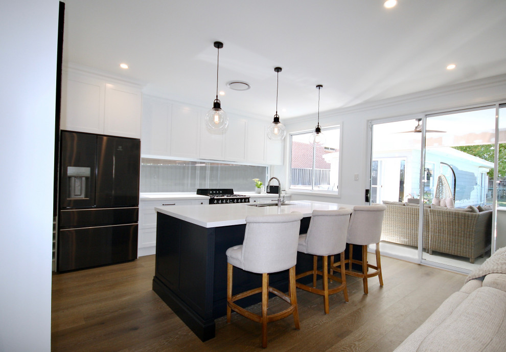 Photo of a mid-sized transitional single-wall open plan kitchen in Sydney with an undermount sink, shaker cabinets, white cabinets, quartz benchtops, window splashback, black appliances, medium hardwood floors, with island, brown floor and white benchtop.
