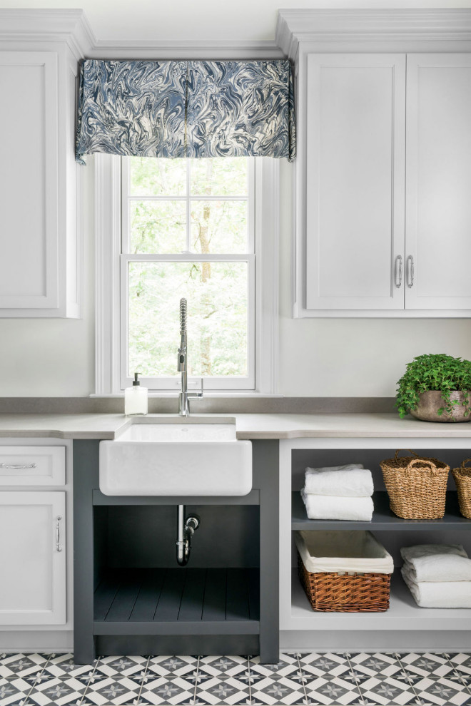 This is an example of a large transitional l-shaped dedicated laundry room in Atlanta with a farmhouse sink, recessed-panel cabinets, white cabinets, quartz benchtops, grey splashback, engineered quartz splashback, white walls, ceramic floors, a side-by-side washer and dryer, multi-coloured floor and grey benchtop.