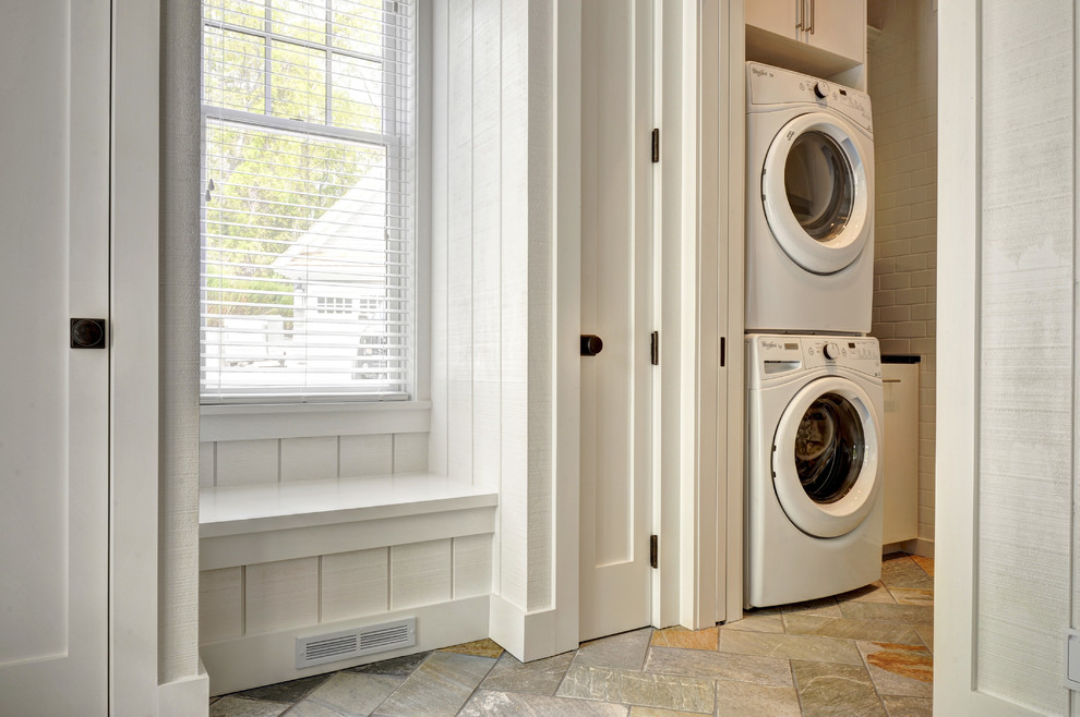 Photo of a mid-sized traditional galley dedicated laundry room in New York with an undermount sink, flat-panel cabinets, white cabinets, soapstone benchtops, white walls, travertine floors and a stacked washer and dryer.
