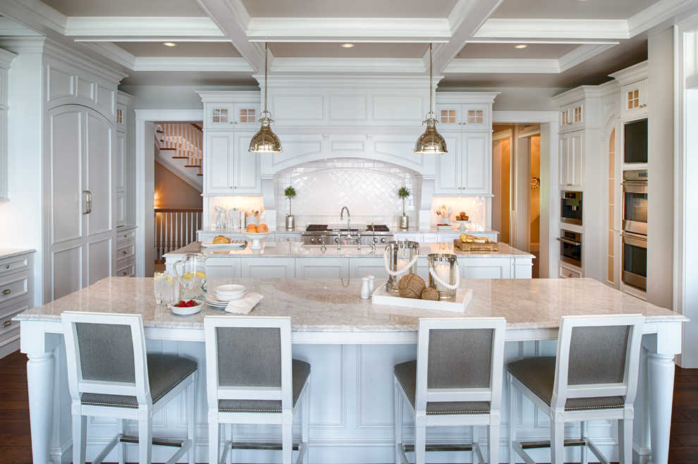 This is an example of an expansive beach style u-shaped eat-in kitchen in Minneapolis with a farmhouse sink, shaker cabinets, white cabinets, marble benchtops, white splashback, porcelain splashback, white appliances, medium hardwood floors, multiple islands and brown floor.