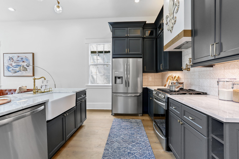 This is an example of a mid-sized arts and crafts l-shaped eat-in kitchen in New Orleans with a farmhouse sink, shaker cabinets, blue cabinets, marble benchtops, beige splashback, ceramic splashback, stainless steel appliances, medium hardwood floors, with island, brown floor and white benchtop.