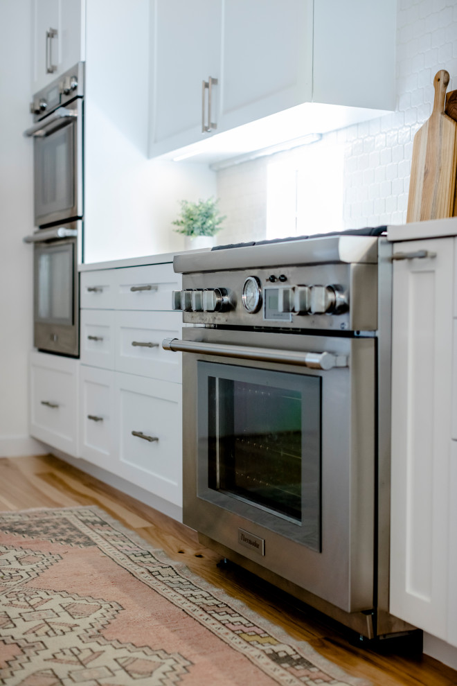 Large transitional l-shaped light wood floor and brown floor eat-in kitchen photo in Austin with a single-bowl sink, shaker cabinets, white cabinets, quartz countertops, white backsplash, ceramic backsplash, stainless steel appliances, an island and white countertops